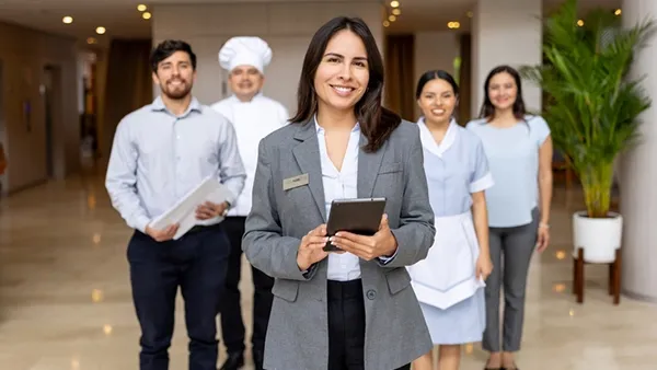 Photo of five hospitality staff standing in a hotel lobby; a person in a gray blazer with a name badge holds a tablet in the foreground, with colleagues behind wearing business attire, a chef’s uniform, and a housekeeping uniform. Large potted plant and softly lit interior in the background.